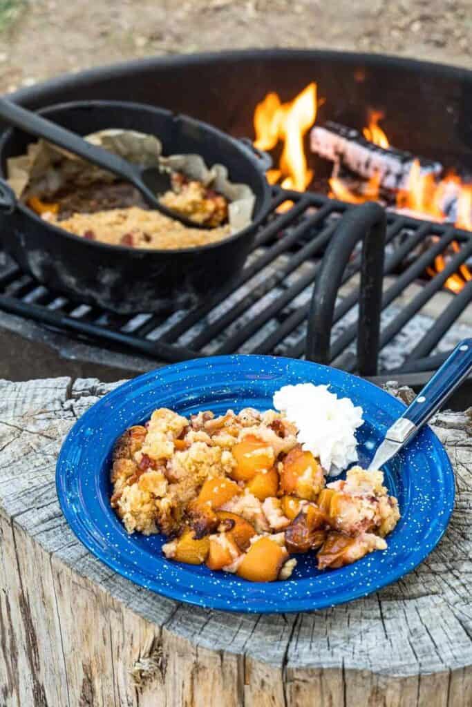 Camp bowl of peach cobbler in front of a lit campfire with a grill. 