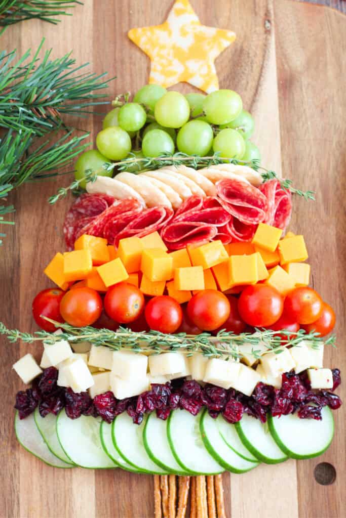 A kid-friendly Christmas tree charcuterie board made with green grapes, crackers, salami, cheddar and white cheese, cherry tomatoes, dried cranberries, cucumber slices, fresh herbs, and a star-shaped cheese topper.