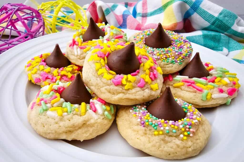 Soft easter blossom cookies topped with colorful spring sprinkles and chocolate kiss candies arranged on a white plate. The festive cookies are decorated in pastel colors for an easter dessert.