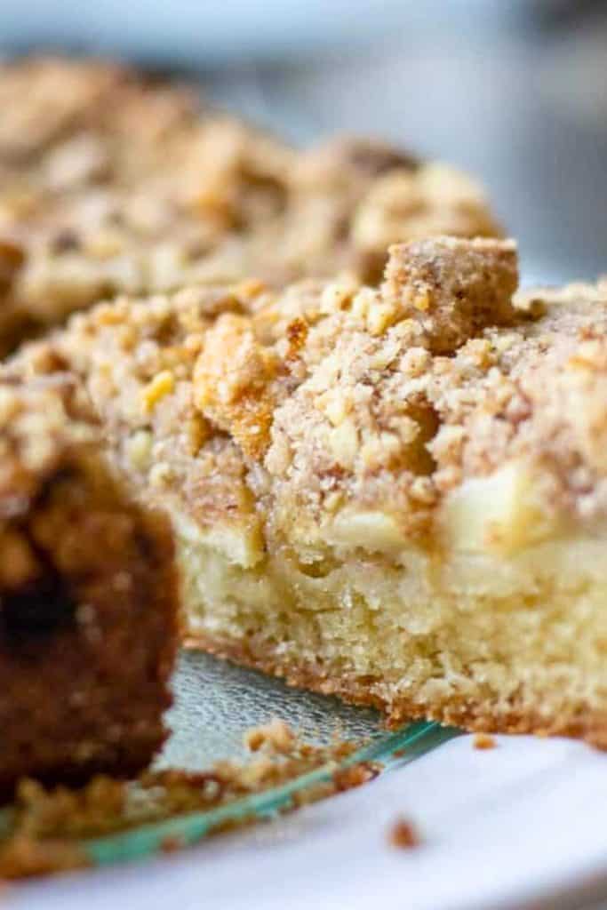 Close-up of a slice of apple crumb cake, showing its soft, buttery interior layered with tender apple pieces and topped with a thick, golden cinnamon streusel. Crumbs are scattered on the plate.