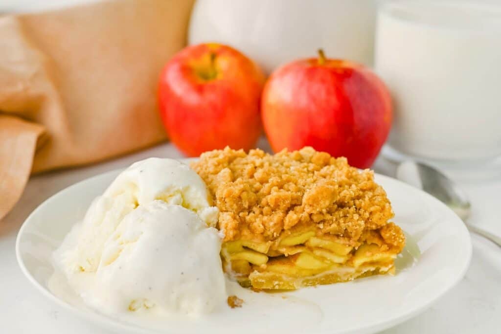 Slice of apple crumble pie bars with layers of cinnamon apples and a golden crumb topping served with melting vanilla ice cream. With two red apples and a glass of milk in the background for an easy apple dessert.