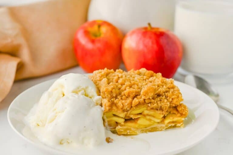 Slice of apple crumble pie bars with layers of cinnamon apples and a golden crumb topping served with melting vanilla ice cream. With two red apples and a glass of milk in the background for an easy apple dessert.