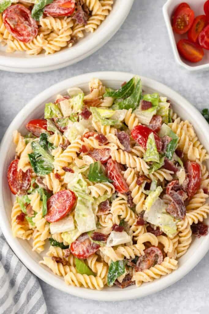 Bowl of easy BLT pasta salad with rotini pasta crispy bacon cherry tomatoes and chopped romaine lettuce tossed in a creamy dressing. The pasta salad combines classic BLT flavors with tender pasta for a hearty cold camping lunch.