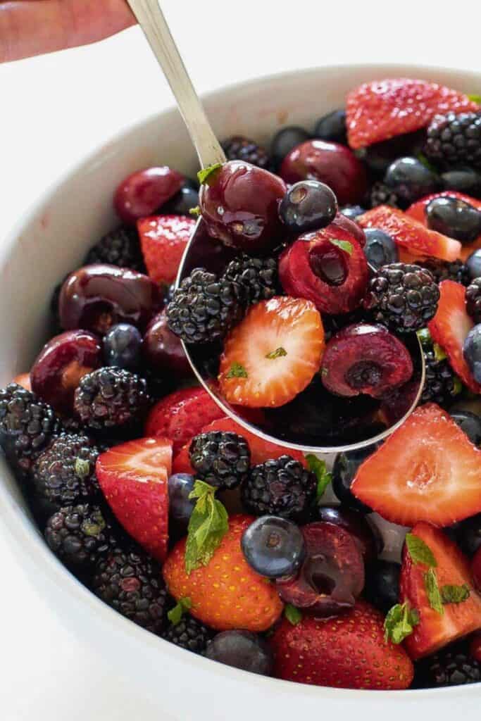 Berry fruit salad in a white bowl filled with fresh strawberries, blueberries, blackberries, and halved cherries, lightly glistening with juice and topped with small mint leaves. A spoon lifts a colorful mix of berries.