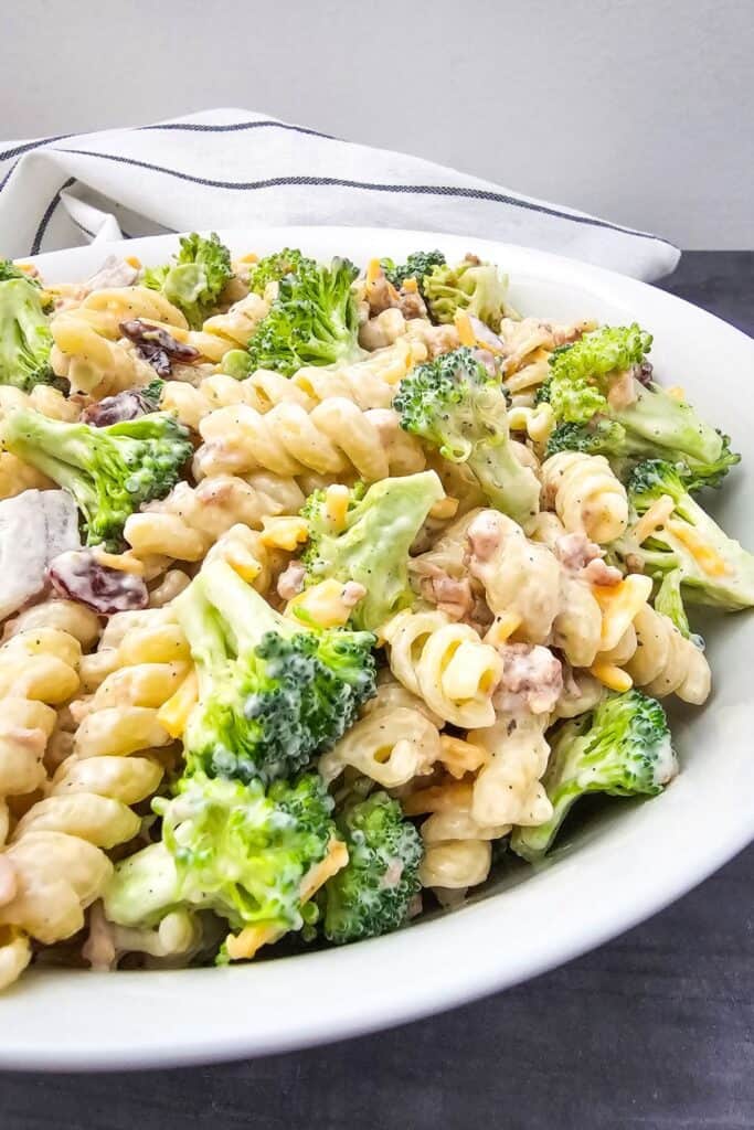 Close-up side view of a creamy broccoli pasta salad in a white bowl, showcasing rotini pasta, fresh broccoli, bacon bits, dried cranberries, cheddar cheese, and chicken pieces, all coated in a rich mayonnaise-based dressing.