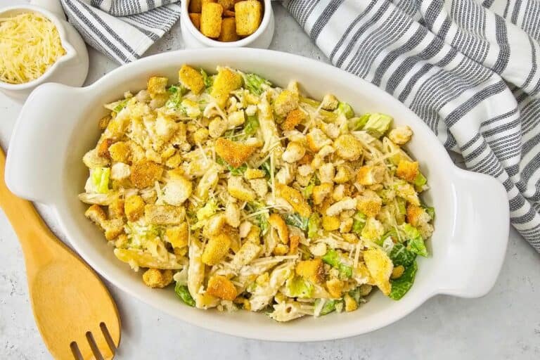 Creamy Caesar pasta salad served in a white oval dish, topped with crunchy croutons, grated Parmesan, and chopped romaine lettuce. Small bowls of extra cheese and croutons sit nearby with a wooden serving spoon and striped cloth, suggesting a fresh, ready to serve side dish.