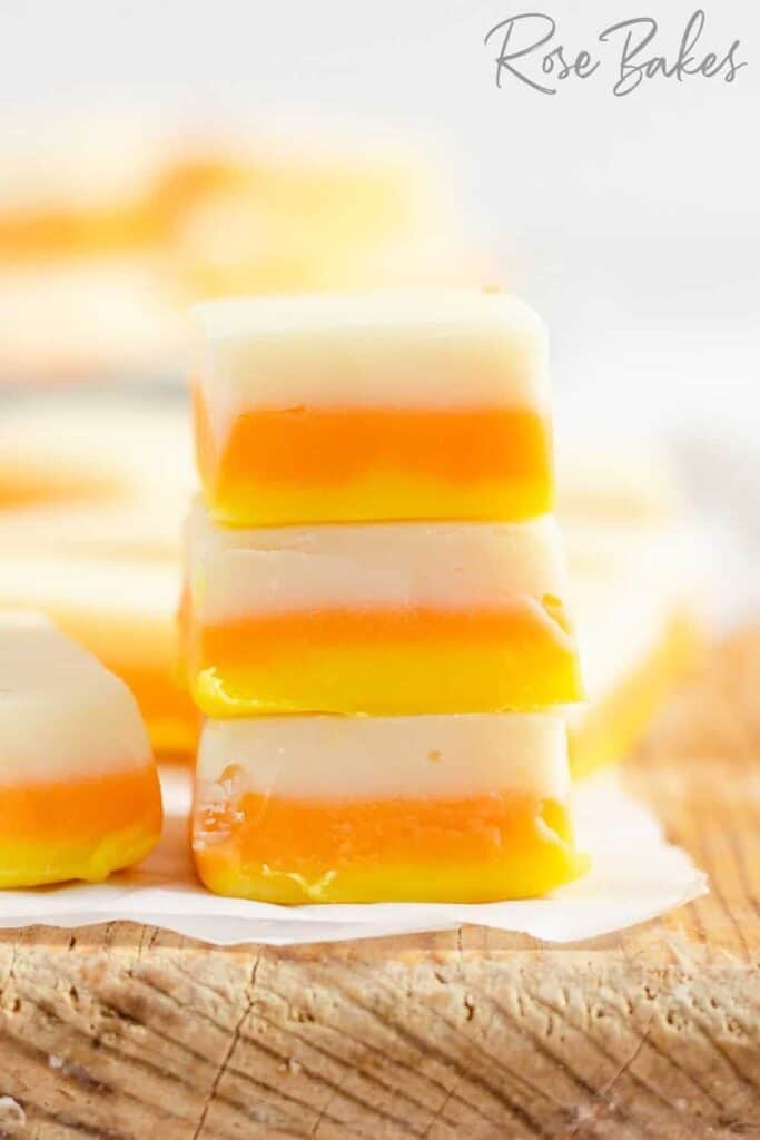 A stack of candy corn inspired fudge squares with distinct white, orange, and yellow layers arranged on a rustic wooden board. The colorful bite sized treats make festive fall desserts for a crowd.