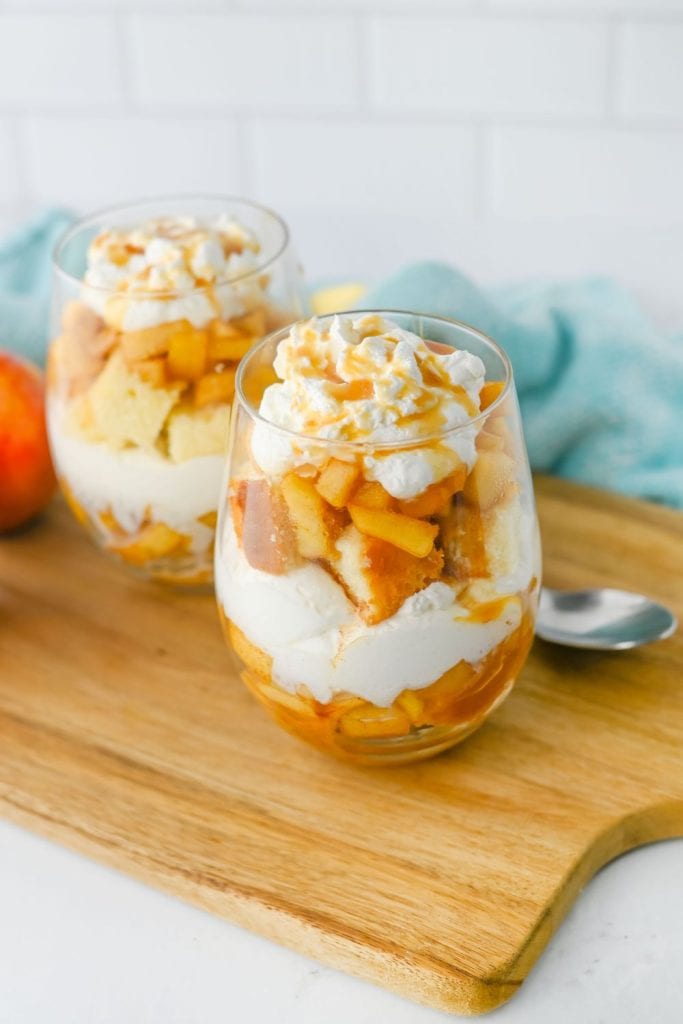 Two finished apple trifles in glass cups are layered with spiced apples, whipped topping, and chunks of pound cake, then topped with a swirl of whipped cream and caramel drizzle. They rest on a wooden board with a spoon and soft blue cloth in the background, ready to serve.