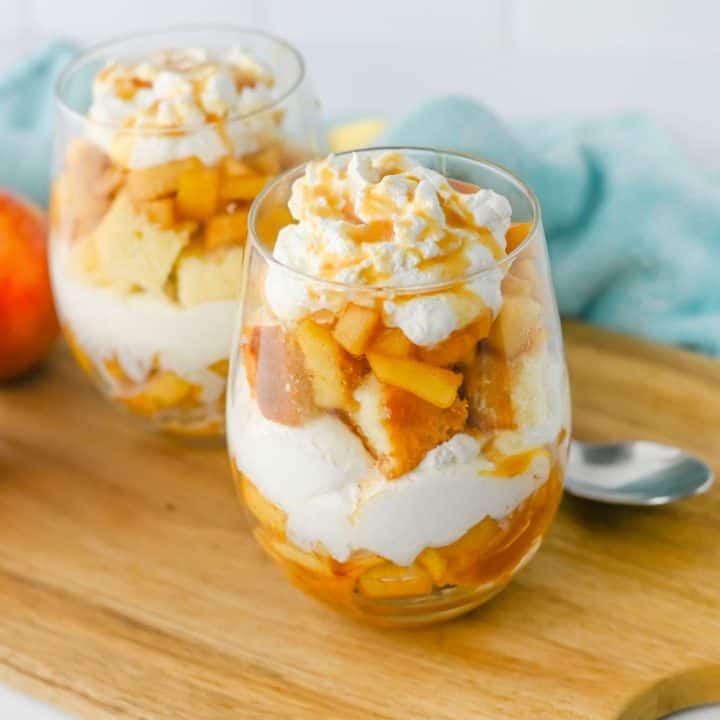 Two finished apple trifles in glass cups are layered with spiced apples, whipped topping, and chunks of pound cake, then topped with a swirl of whipped cream and caramel drizzle. They rest on a wooden board with a spoon and soft blue cloth in the background, ready to serve.