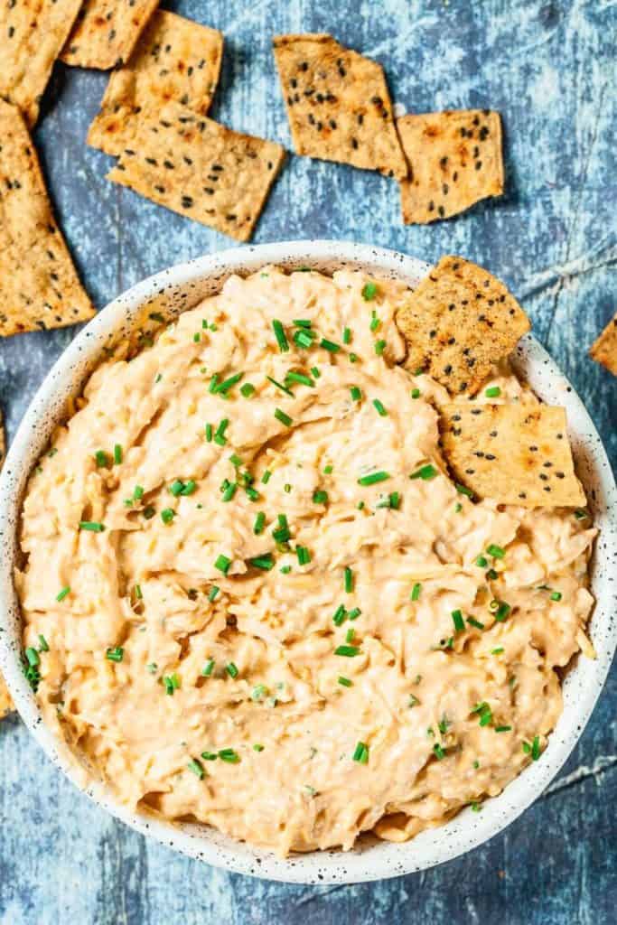 Easy Cheesy Chicken Dip served in a speckled white bowl, topped with chopped chives for garnish. Surrounded by seeded crackers, this creamy cold game day dip is loaded with shredded chicken and melted cheese for a hearty snack.
