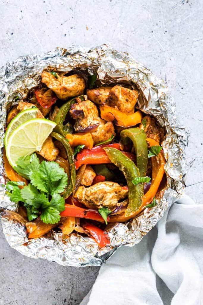 An overhead view of a  round tinfoil pack with chicken fajita ingredients inside, topped with lemon slices and cilantro garnish. 