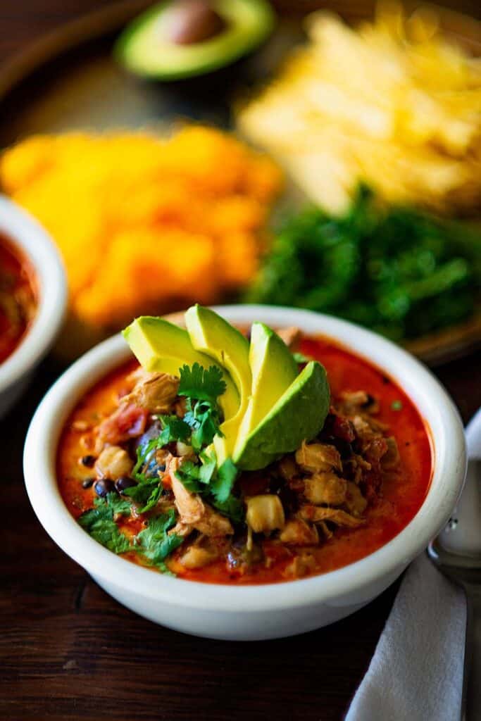 Small ceramic bowl of tomato based chicken tortilla soup with beans, and topped with avocado slices fanned out. 