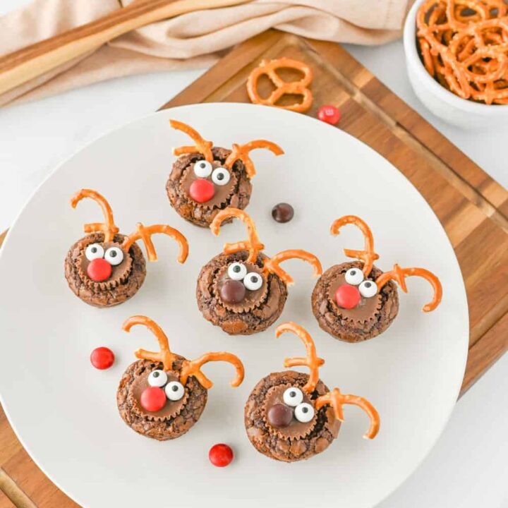 White plate of easy Christmas snacks for kids featuring reindeer brownie bites with candy eyes, pretzel antlers, and M&M noses. The festive treats are displayed on a wooden board with scattered candies, pretzels, and a wooden spoon in the background.