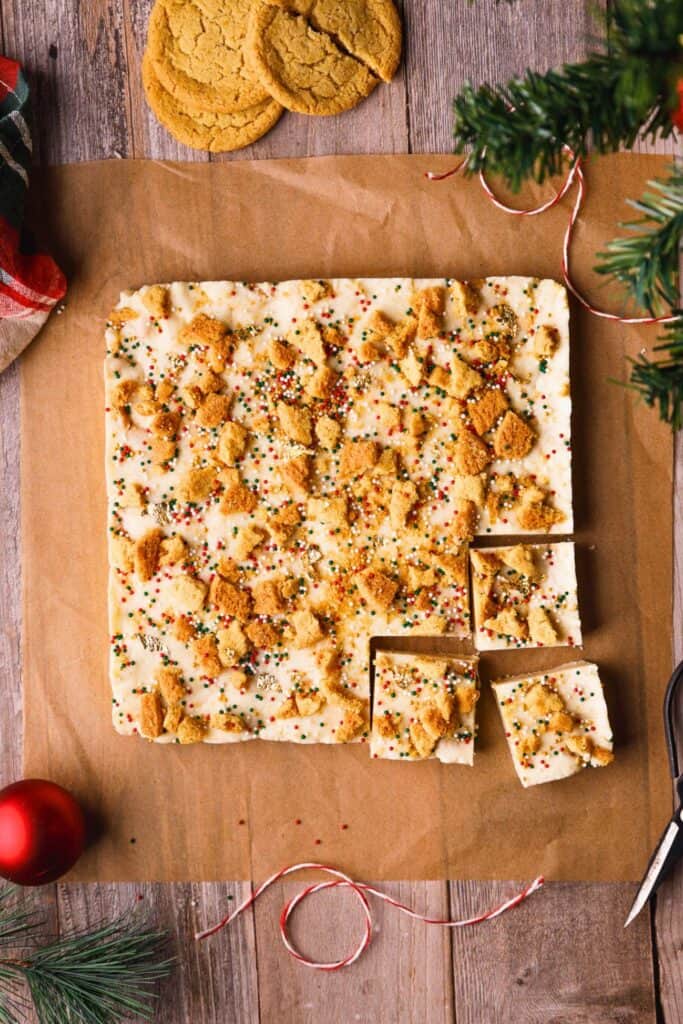 Sugar cookie fudge topped with crushed sugar cookies and festive red, green, and white sprinkles, cut into squares on parchment paper. A few whole cookies, ribbon, and pine branches add a cozy Christmas touch around the edges.