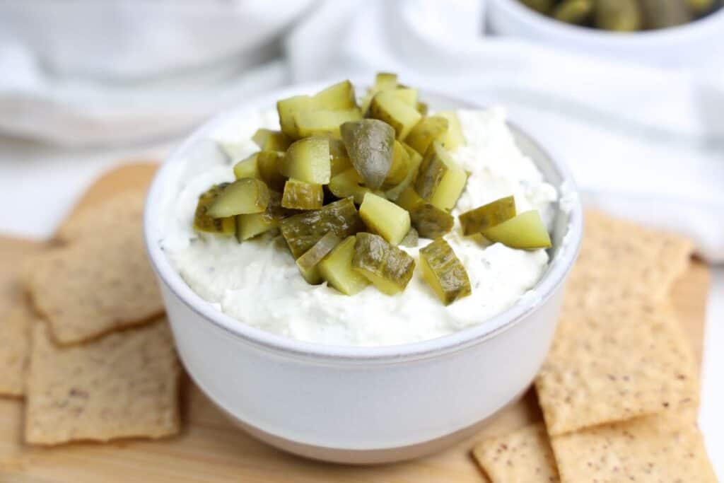 A white bowl filled with creamy dill pickle dip, topped with chopped pickles, sits on a wooden board surrounded by whole grain crackers. The dip has a thick, whipped texture with visible ranch flavored herb flecks.