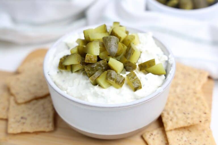A white bowl filled with creamy dill pickle dip, topped with chopped pickles, sits on a wooden board surrounded by whole grain crackers. The dip has a thick, whipped texture with visible ranch flavored herb flecks.