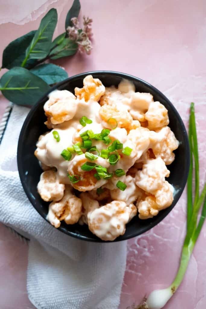 Creamy coconut shrimp coated in a rich white sauce are piled in a black bowl and topped with chopped green onions. The bowl rests on a folded napkin with fresh green onions and decorative greenery arranged on the pink surface around it.