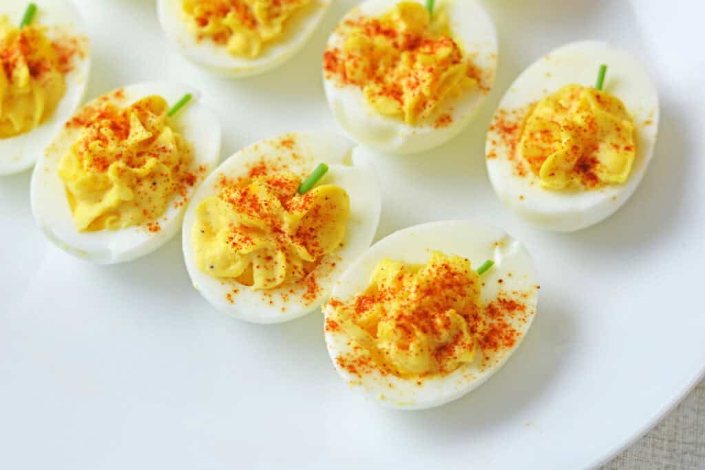 Overhead close up view of pumpkin shaped deviled eggs with creamy yellow filling dusted with smoked paprika and topped with green chive stems on a plate. These festive bites are easy Fall and Halloween appetizers designed to resemble miniature pumpkins.