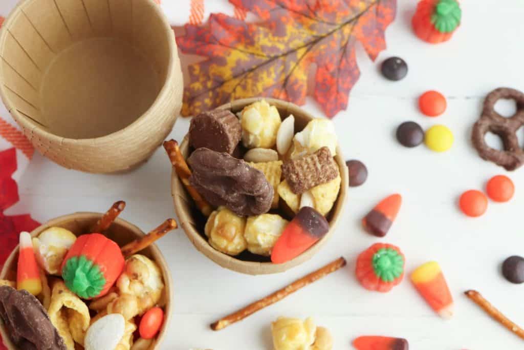 Overhead view of paper cups filled with fall Chex mix snack, including chocolate-covered pretzels, caramel popcorn, Chex cereal, candy corn, peanuts, pretzel sticks, and candy pumpkins, surrounded by scattered fall candies and decorative leaves on a white table.