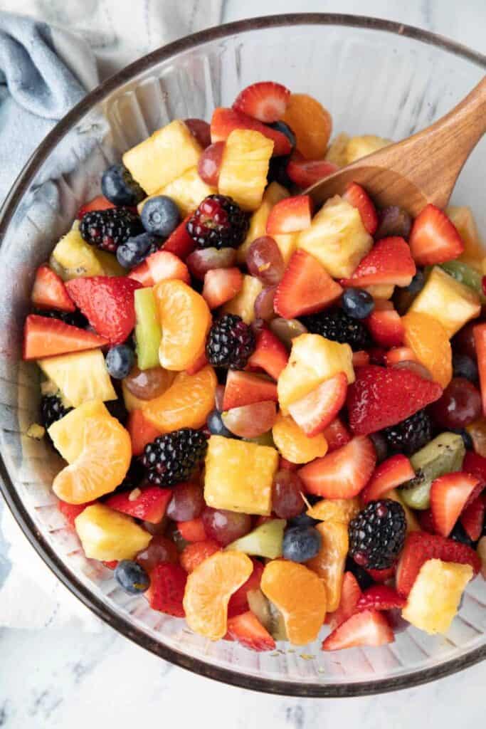 A colorful fruit salad in a clear glass bowl, filled with a mix of strawberries, blueberries, blackberries, pineapple chunks, mandarin orange segments, red grapes, and kiwi slices. A wooden spoon rests inside the bowl, ready for serving.