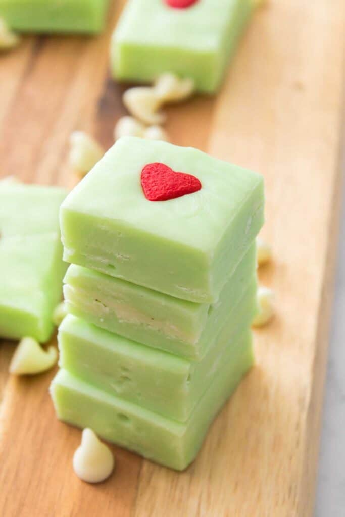 Stacked squares of creamy green Grinch fudge, each topped with a small red heart sprinkle. The fudge rests on a wooden board with scattered white chocolate chips, giving a playful nod to the classic holiday story.