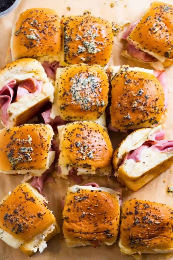 Tray of mini ham and cheese sliders made with golden slider buns, layered with slices of ham and melted cheese, and topped with a buttery poppy seed glaze. These warm, pull-apart tailgate snacks are arranged on parchment paper for easy serving.
