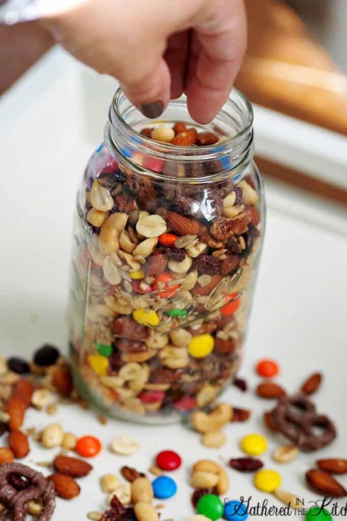 Hand reaching into a mason jar filled with salty and sweet travel trail mix with muts, and M&Ms. 