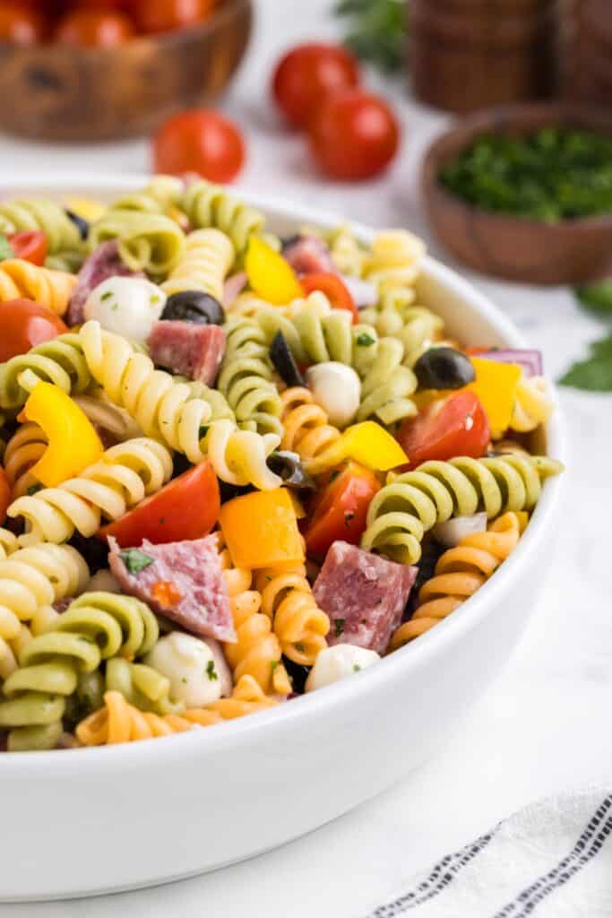 Easy Italian pasta salad with tri color rotini tossed with cherry tomatoes, mozzarella balls, salami, olives, and yellow bell peppers. The colorful pasta salad is mixed with herbs and served in a large white bowl with fresh ingredients blurred in the background.