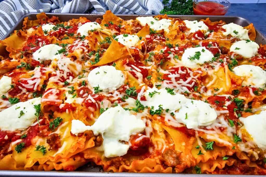 Easy weeknight meal of sheet pan lasagna with ruffled noodles coated in tomato meat sauce and melted mozzarella. Creamy dollops of ricotta and fresh parsley are scattered across the top.