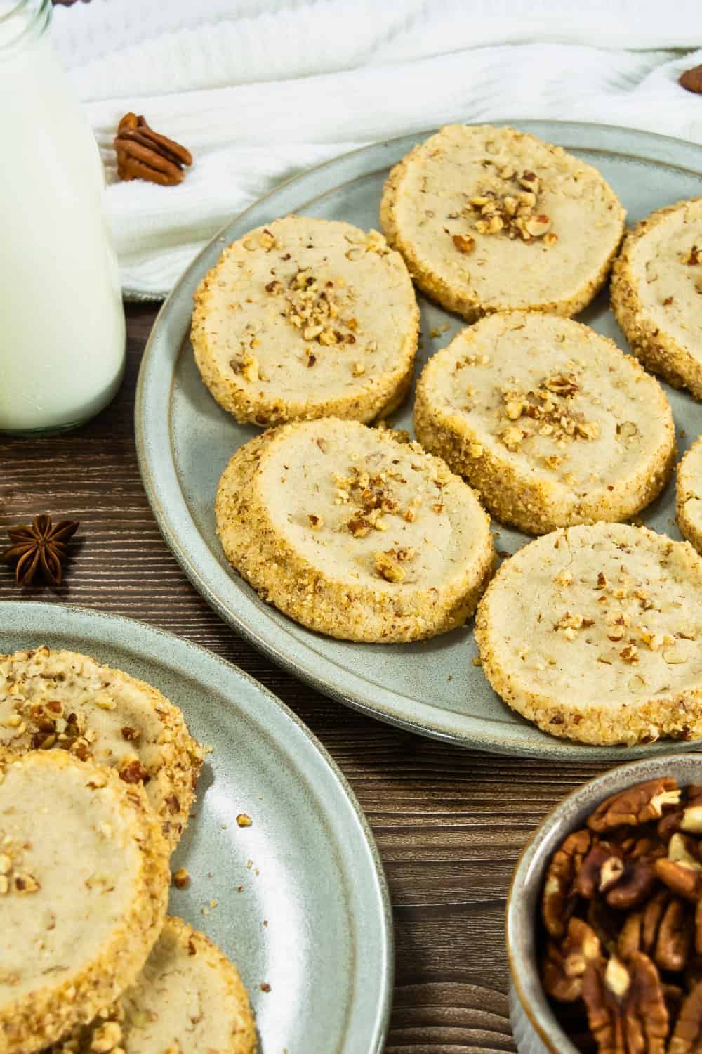 Pecan shortbread cookies with chopped pecans baked throughout and around the edges, arranged on ceramic plates beside a bowl of pecans and a bottle of milk.