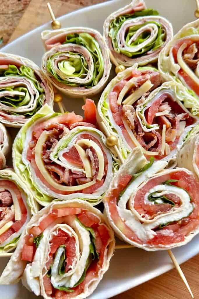 Three rows of pinwheel sandwiches on a plate. Includes ham and cheese, caprese (basil, tomato, mozzarella) and turkey tomato, cheese, bacon and lettuce pinwheels wrapped in soft tortillas.
