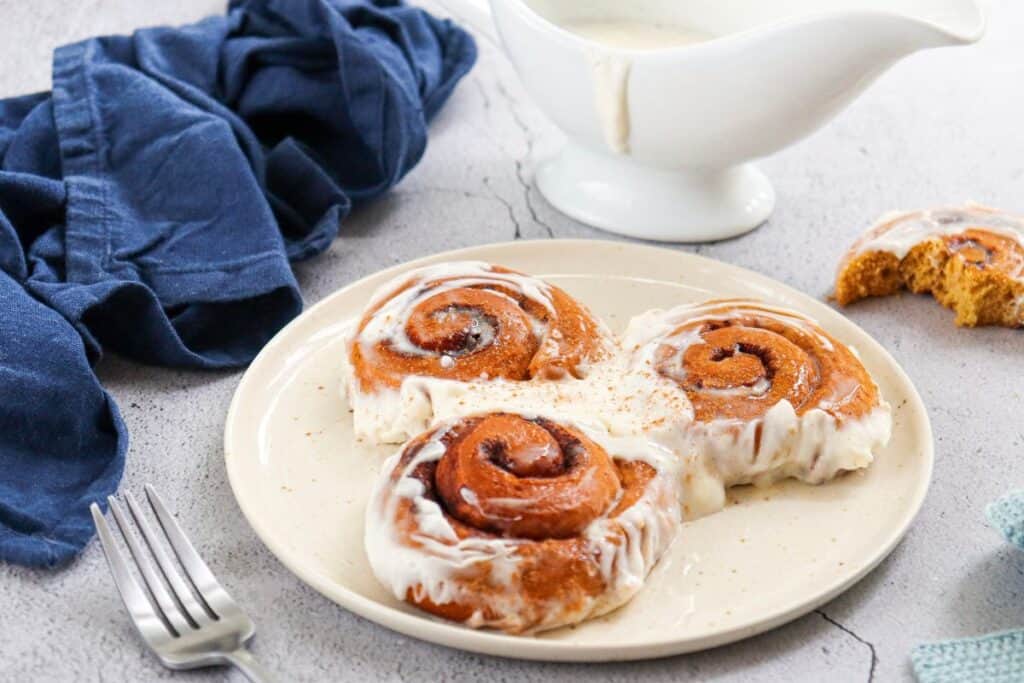 Plate of three pumpkin cinnamon rolls with Maple Cream Cheese Icing. beside a tea towel and fork.