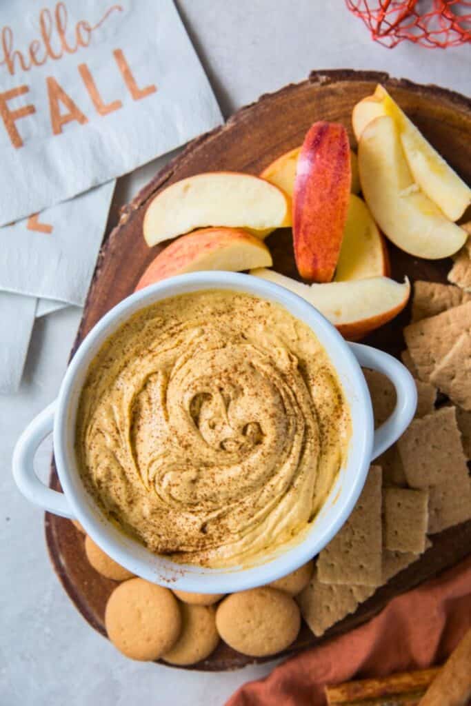 Easy pumpkin cheesecake dip swirled in a white bowl and dusted with warm spices. Sliced apples, graham crackers, and small cookies surround the creamy dip on a rustic wooden serving board beside a napkin that reads "hello FALL".