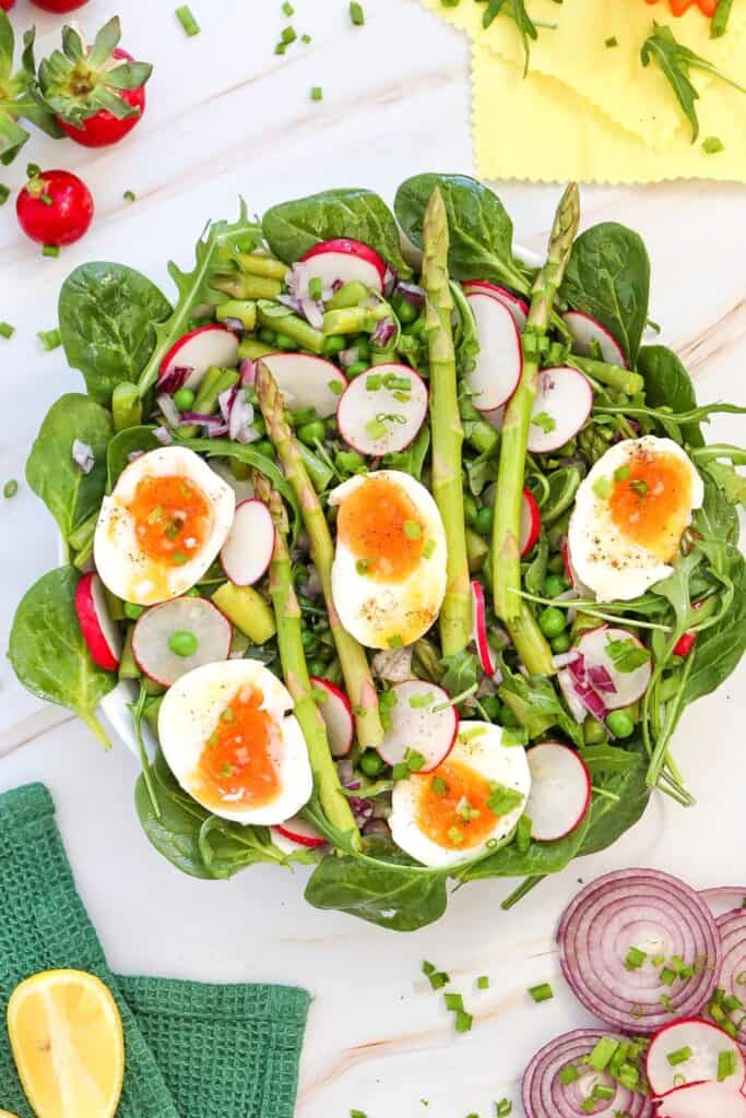 Overhead voew of plate of quick asparagus side salad topped with halved boiled eggs, beside a green tea towl, half a red onion and cut lemons. 