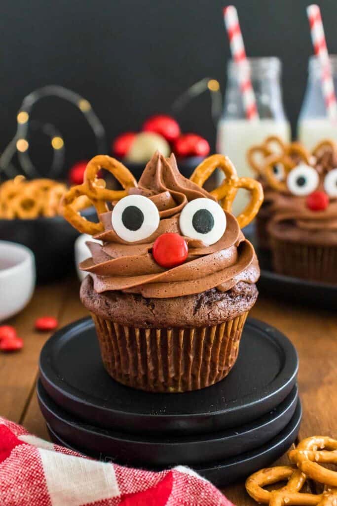 A chocolate cupcake is decorated with swirls of chocolate frosting, candy eyes, a red M&M nose, and pretzels for antlers to resemble a reindeer. This fun holiday dessert is part of a festive setup with milk bottles, ornaments, and more easy reindeer cupcakes for kids in the background.