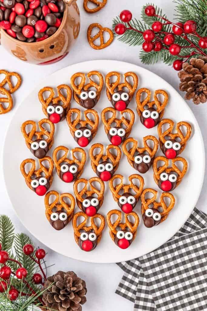 A white plate is filled with reindeer Rolo pretzel Christmas treats made from mini pretzels for antlers, chocolate Rolos as faces, candy eyes, and red or brown M&M noses. The festive setup is surrounded by pinecones, holiday greenery, and a bowl of red and brown M&Ms.