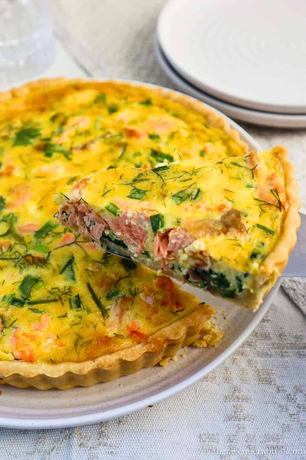 A golden Easy Salmon Spinach Quiche filled with chunks of salmon, spinach, green onions, and fresh dill in a flaky pastry crust, with one slice lifted from the quiche to show the creamy filling.