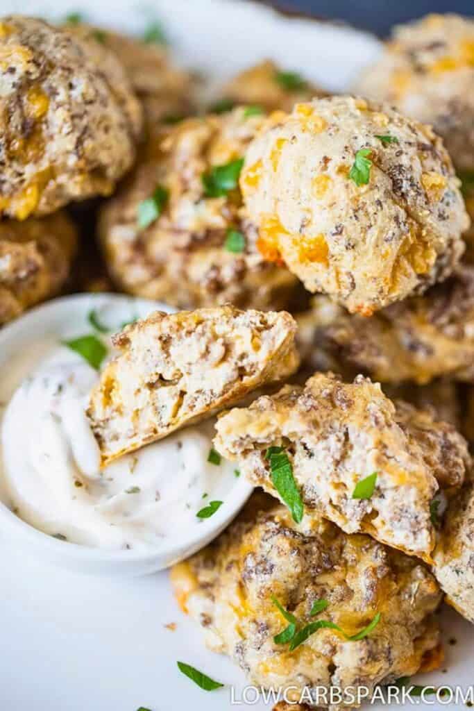 Close-up of easy sausage meatballs baked until golden brown, with melted cheese visible throughout and fresh herbs sprinkled on top. One meatball is cut open to show the juicy, tender interior, served alongside a small dish of creamy dipping sauce.