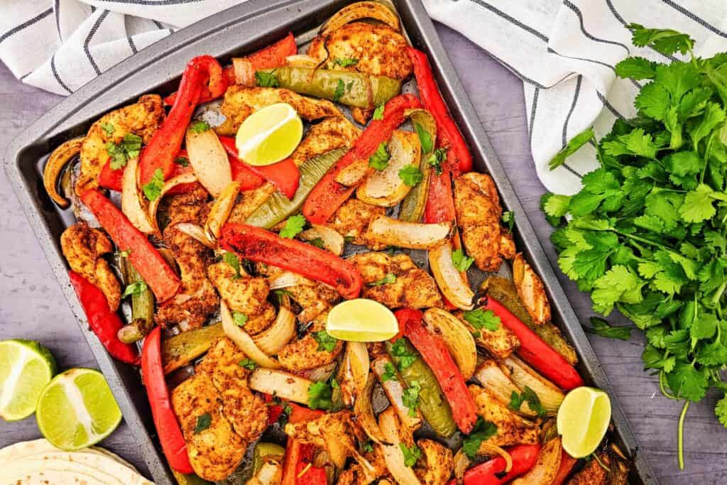 Sheet pan chicken fajitas with seasoned sliced chicken breast roasted alongside red and green bell peppers and onion wedges, topped with fresh cilantro and lime wedges. The colorful one pan chicken fajitas are served straight from the baking tray for an easy weeknight dinner with tortillas on the side.