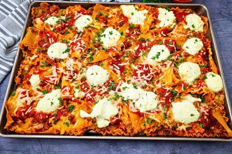 Overhead view of a layerless sheet pan lasagna with ruffled lasagna noodles covered in rich tomato meat sauce. Dollops of ricotta and melted mozzarella are scattered across the top with fresh parsley.