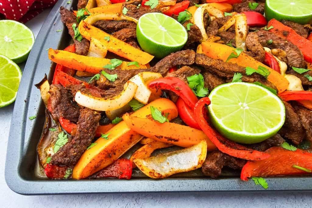 Seasoned strips of steak are roasted on a sheet pan with sliced red and orange bell peppers and onions. Fresh cilantro is scattered over the steak fajitas with bright green lime halves placed on top.