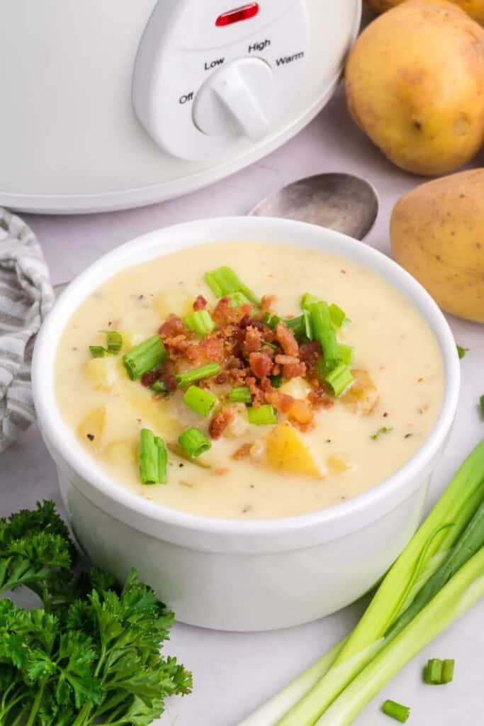 Easy slowcooker potato soup with tender potato chunks is served in a white bowl. Crispy bacon and chopped green onions top the creamy soup with the slow cooker visible in the background.