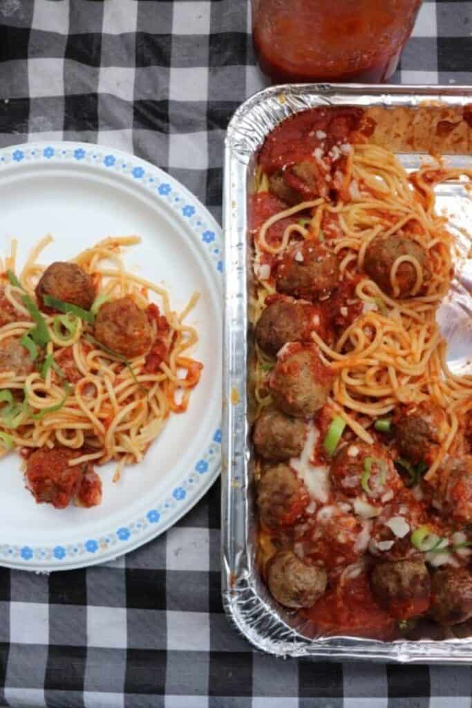 Oerhead view of a picnic tablecloth with a tinfoil pan of meatball spaghetti beside a paper plate with with a pile of the spaghetti.