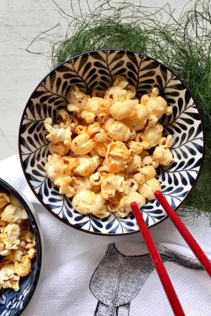 Bowl of easy spicy popcorn seasoned with chili flakes and spices, served in a patterned black and white dish with red chopsticks. The popcorn is lightly golden for a bold, savory snack presentation.