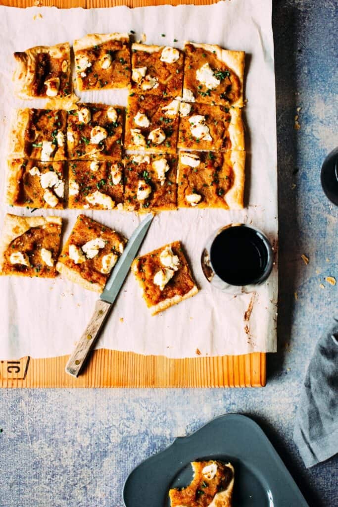 A squash, roasted garlic and goat cheese tart is cut into small squares on parchment paper, with several pieces separated for serving. The golden puff pastry is topped with creamy squash, dollops of goat cheese, and chopped fresh herbs, with a knife and glasses of red wine beside it.
