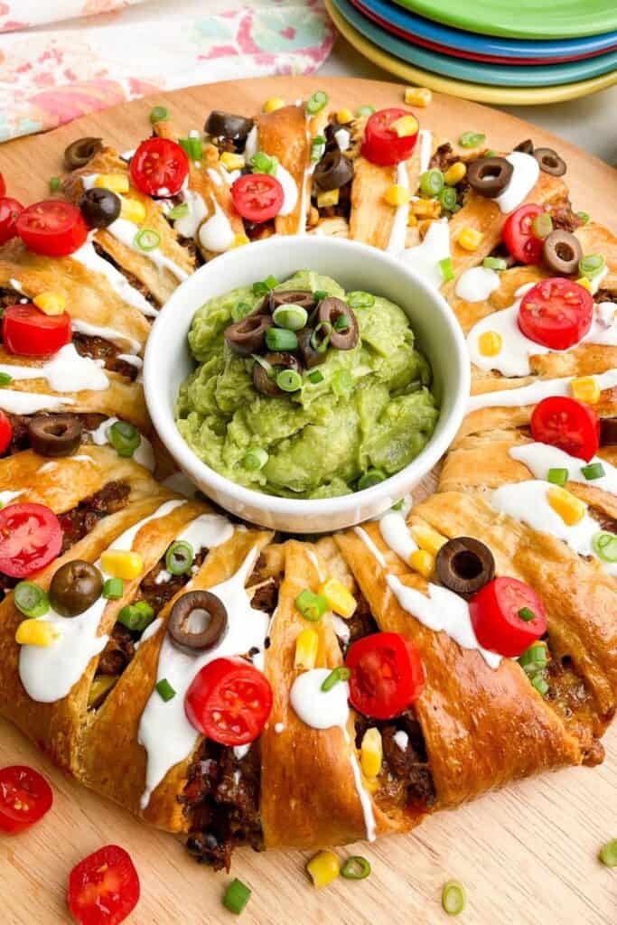 A golden-brown taco ring made with crescent dough, filled with seasoned beef and topped with sour cream, sliced cherry tomatoes, olives, corn, and green onions, arranged around a bowl of guacamole garnished with more olives and scallions—served on a wooden board as an appetizer.