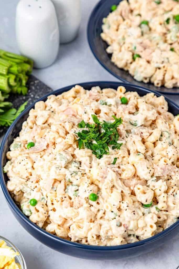 Easy tuna macaroni salad served in a dark blue bowl, featuring elbow macaroni mixed with flaked tuna, green peas, diced celery, and a creamy mayonnaise-based dressing. The dish is topped with cracked black pepper and a garnish of fresh parsley for a simple and classic presentation.