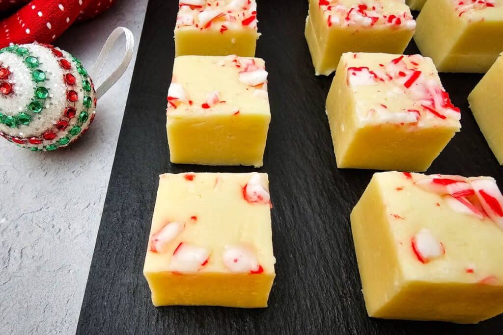 Close-up of white chocolate peppermint fudge squares topped with crushed candy canes on a black serving tray. The creamy fudge has a festive look, complemented by a sparkly red, green, and white ornament beside it,