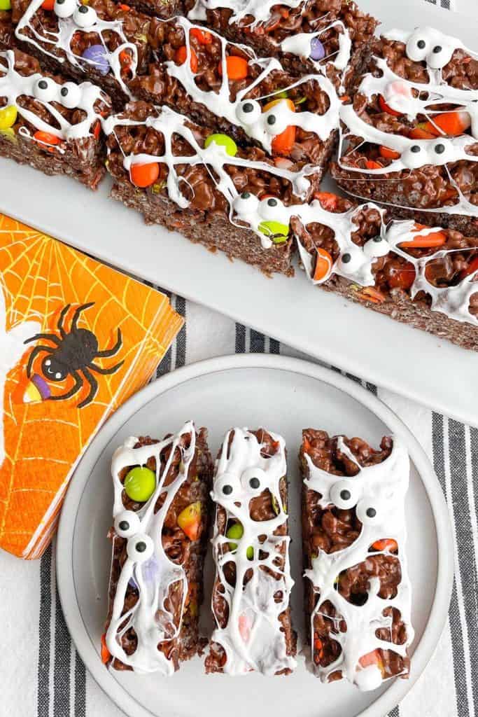 Chocolate crispy bars decorated for Halloween with candy eyes, colorful candy-coated chocolates, and drizzles of white marshmallow to resemble mummies, served on a plate and platter beside an orange napkin with a spiderweb design.