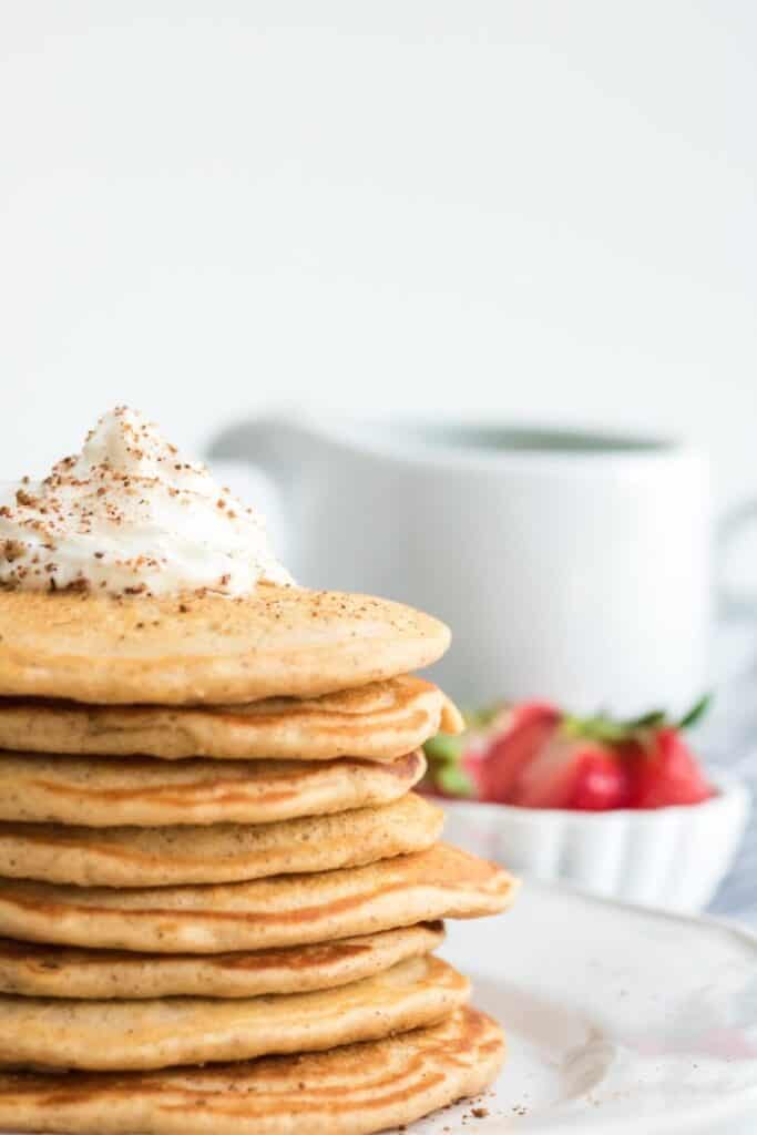 Stack of eight eggnog pancakes topped wth a dollop of whip cream, on a plate. 